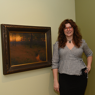 Woman standing in front of a painting showing a landscape, sunset, and owl.