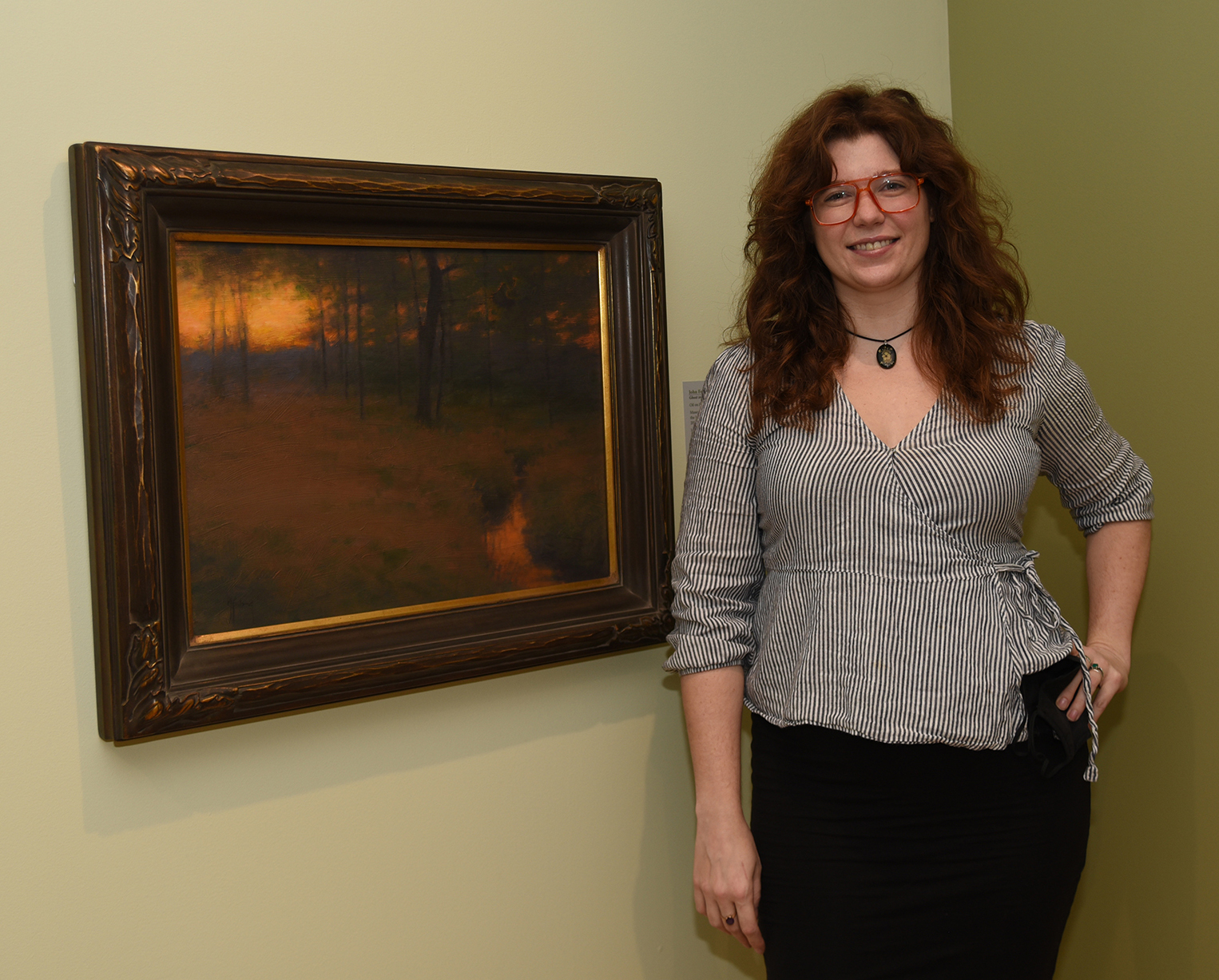 Woman stands next to a painting of a landscape at twilight.