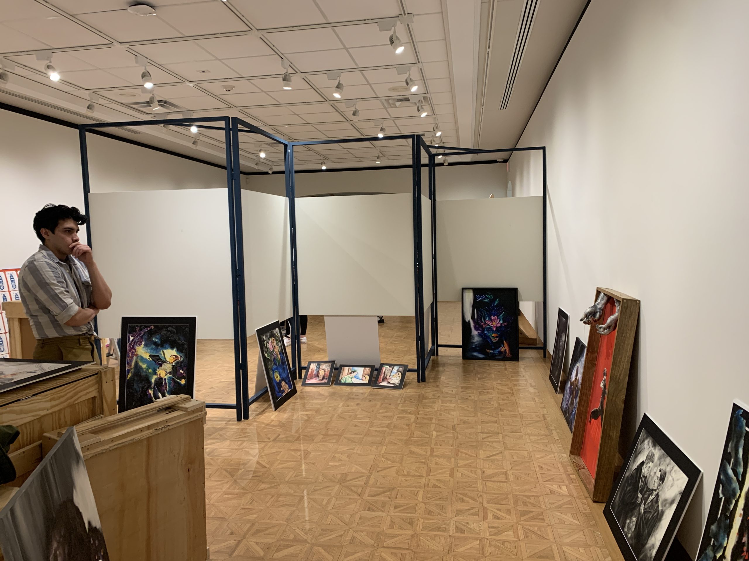 A high school student stands with a hand over his mouth, contemplating the paintings that surround him on the floor. He is preparing to place and hang artwork in the gallery space.