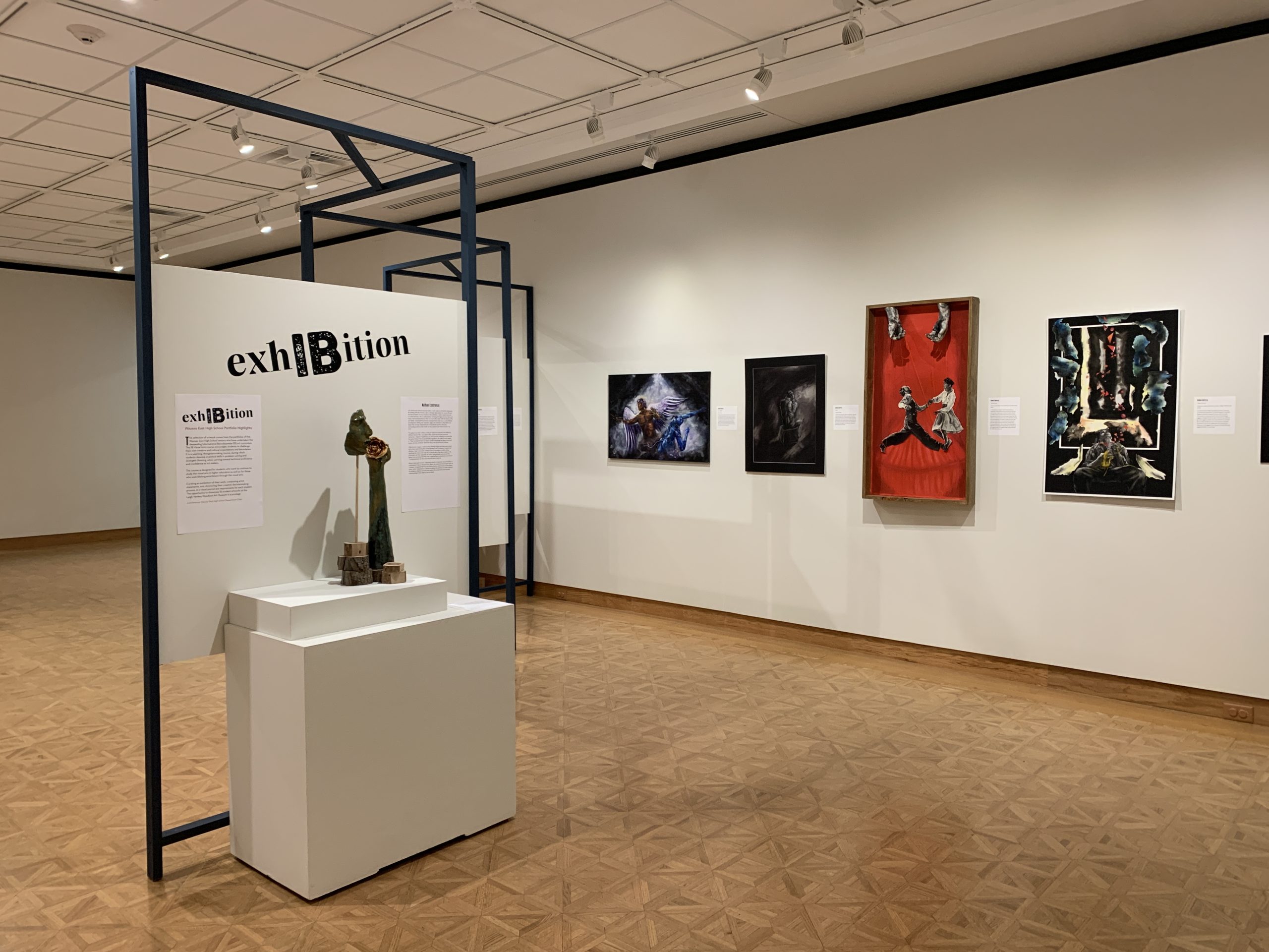 A three dimensional mixed media artwork depicting a face and hand holding a flower sits atop a pedestal in the gallery. Near the sculptural work are four artworks hanging on the wall. One features a dancing couple on red background. Above the couple are two hands holding strings, as if controlling the people dancing like puppets.