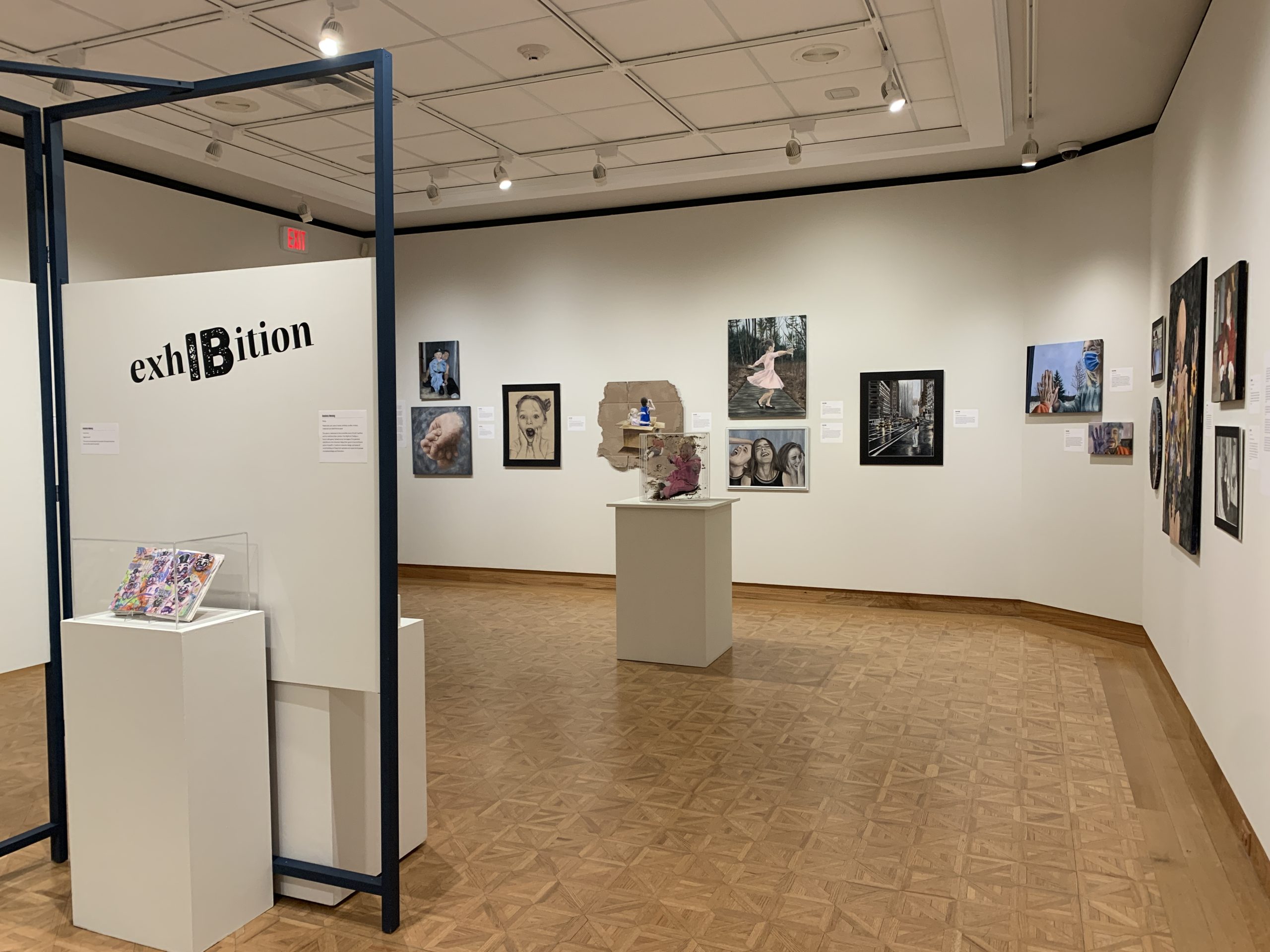 Installation view of the lower level gallery featuring Wausau East High School International Baccalaureate student work. Paintings and drawings featuring an array of figural work hang on the gallery walls. Two pedestals feature three-dimensional work in the space. 
