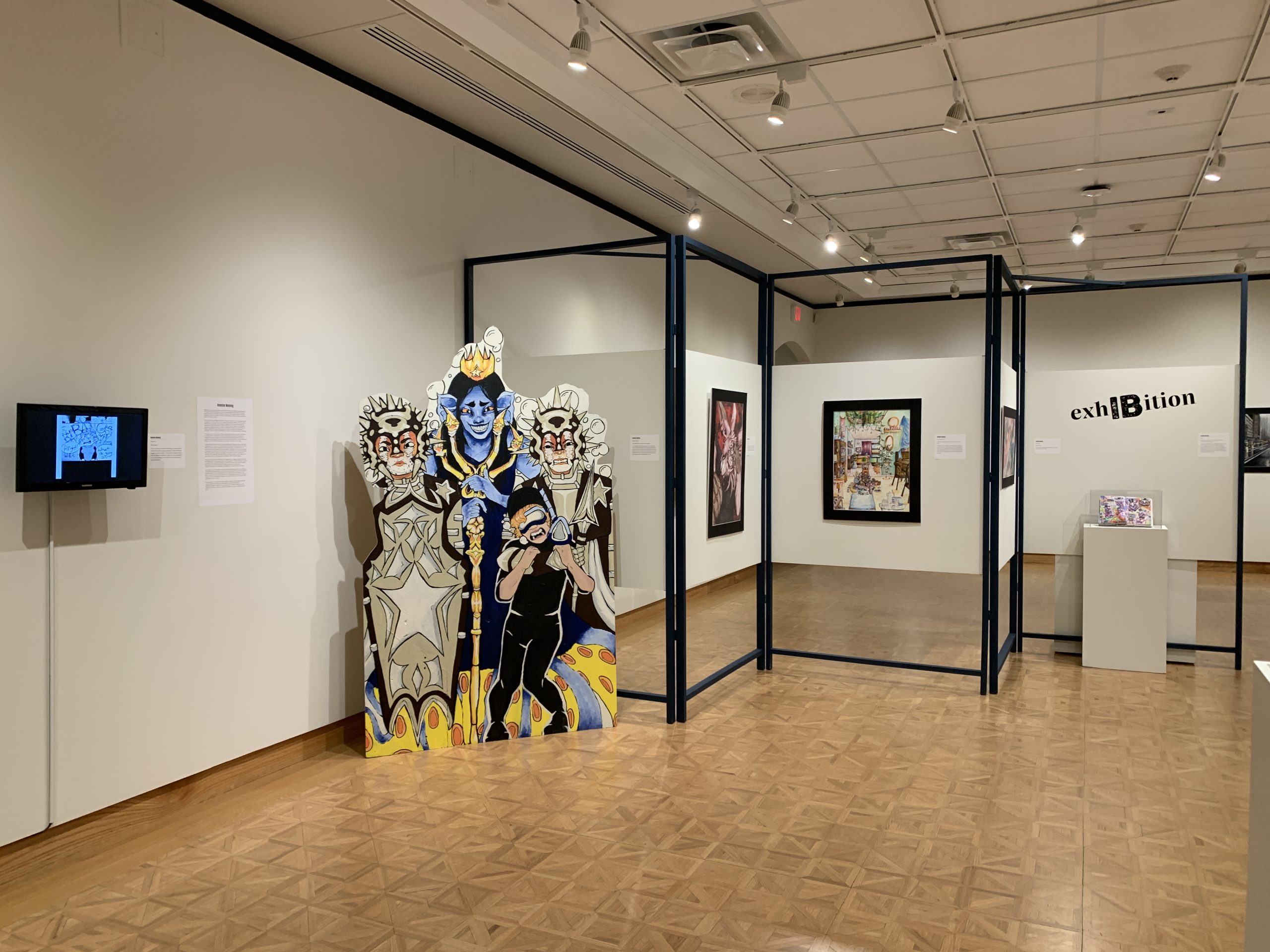 At the center of the image is a large scale, free-standing painting on plywood with three figures in detailed costuming. To the left of the painting hangs a TV screen, scrolling through an illustrated comic. On the other side of the large painting is a movable wall in a zig-zag pattern displaying other two-dimensional work. 