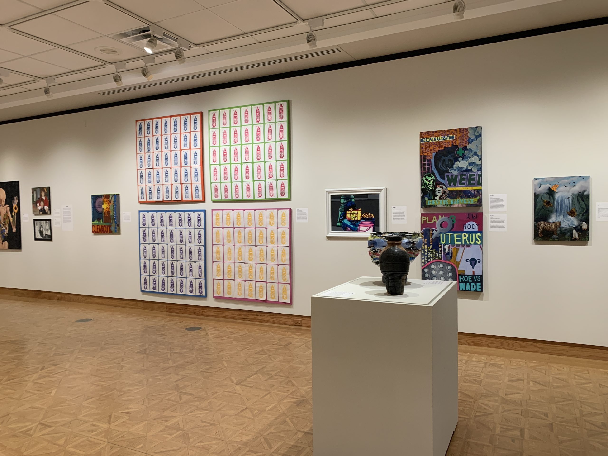 Four large, colorful panels hang on the wall in the center of the image. Each panel contains 30 prints of water bottles. The panels are flanked by paintings on either side and a pedestal holds two three-dimensional artworks in the foreground of the image.