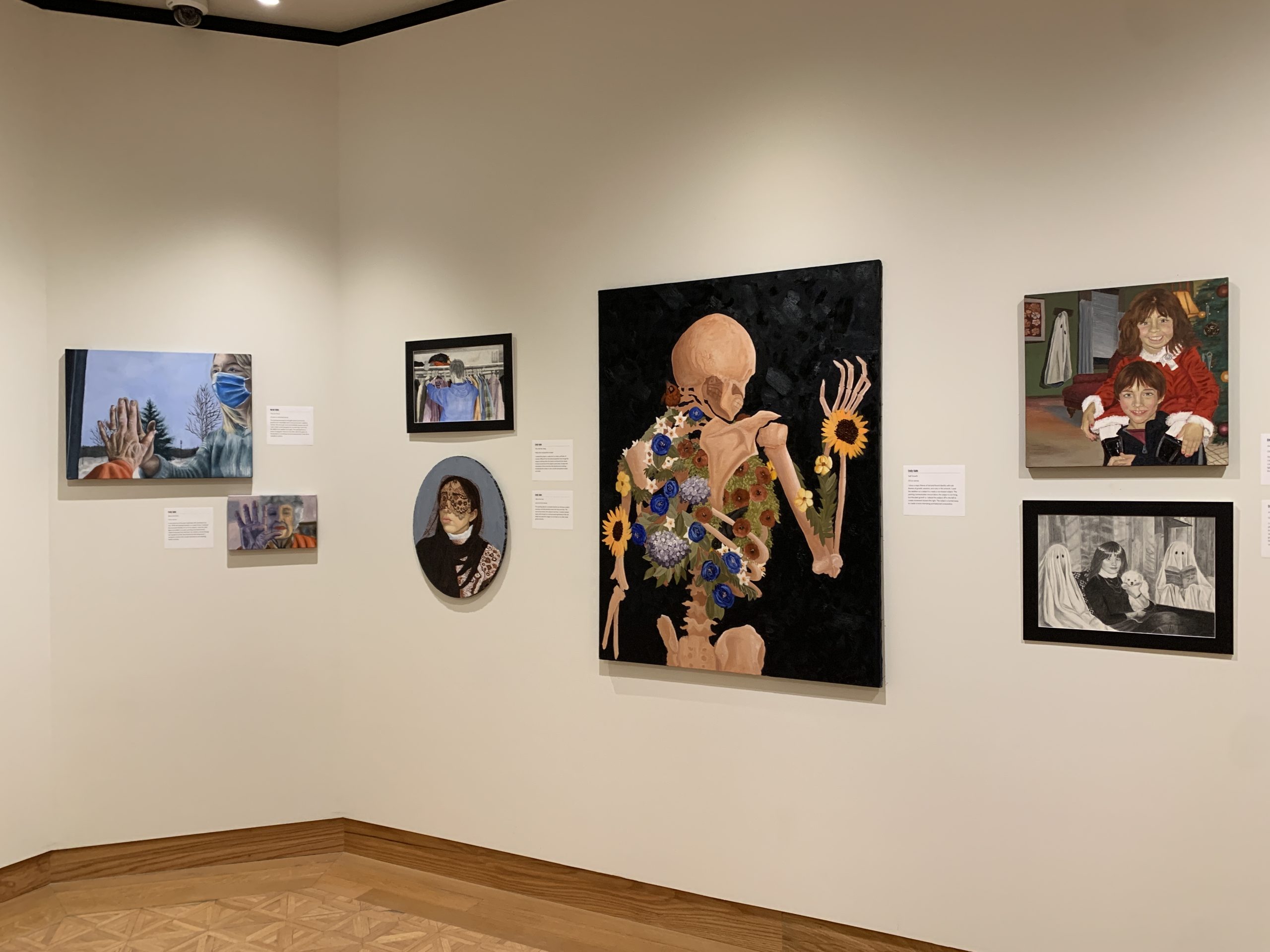 Seven paintings showing various figures and portraits hang on the gallery wall. The largest painting depicts a skeleton figure on a black background facing away from the viewer. The skeleton holds up its right hand and flowers adorn its bones.