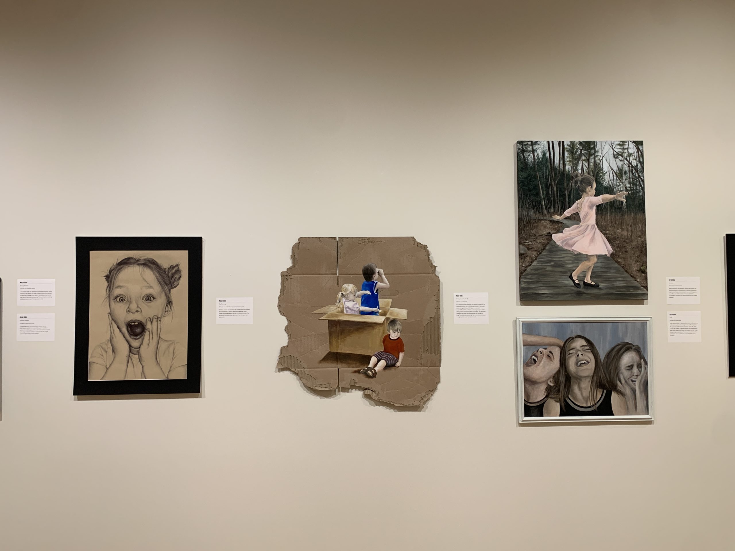 Four drawings and paintings depicting children hang on the gallery wall. A monochromatic drawing of a young girl with a surprised expression hangs to the left of a painting created on cardboard depicting three children playing in a moving box.