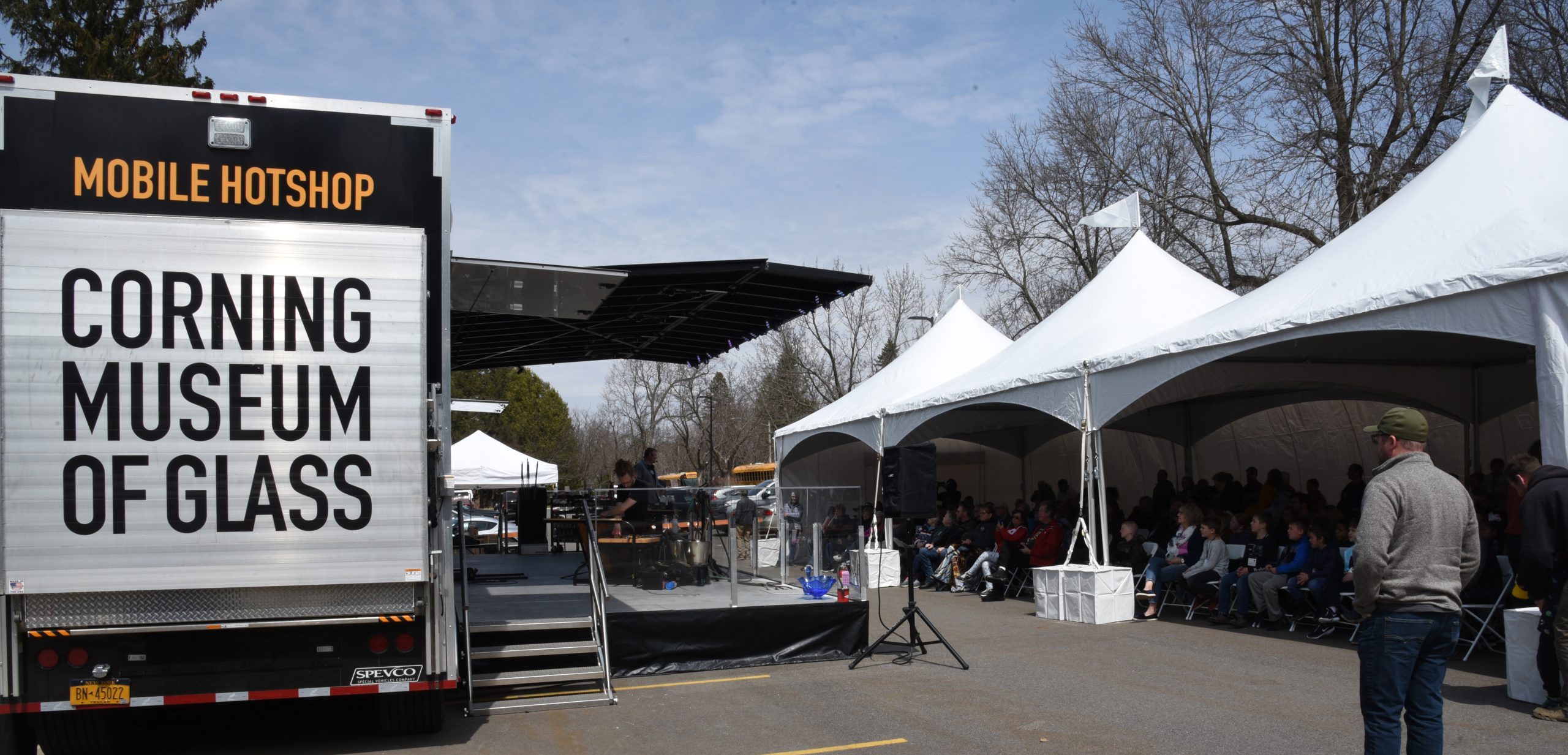 Dozens of people seated beneath a tent listen and watch to glassmaking demonstrations on an outdoor stage that folds out from a semi-tractor trailer that traveled from Corning, N.Y., to Wausau, WI.