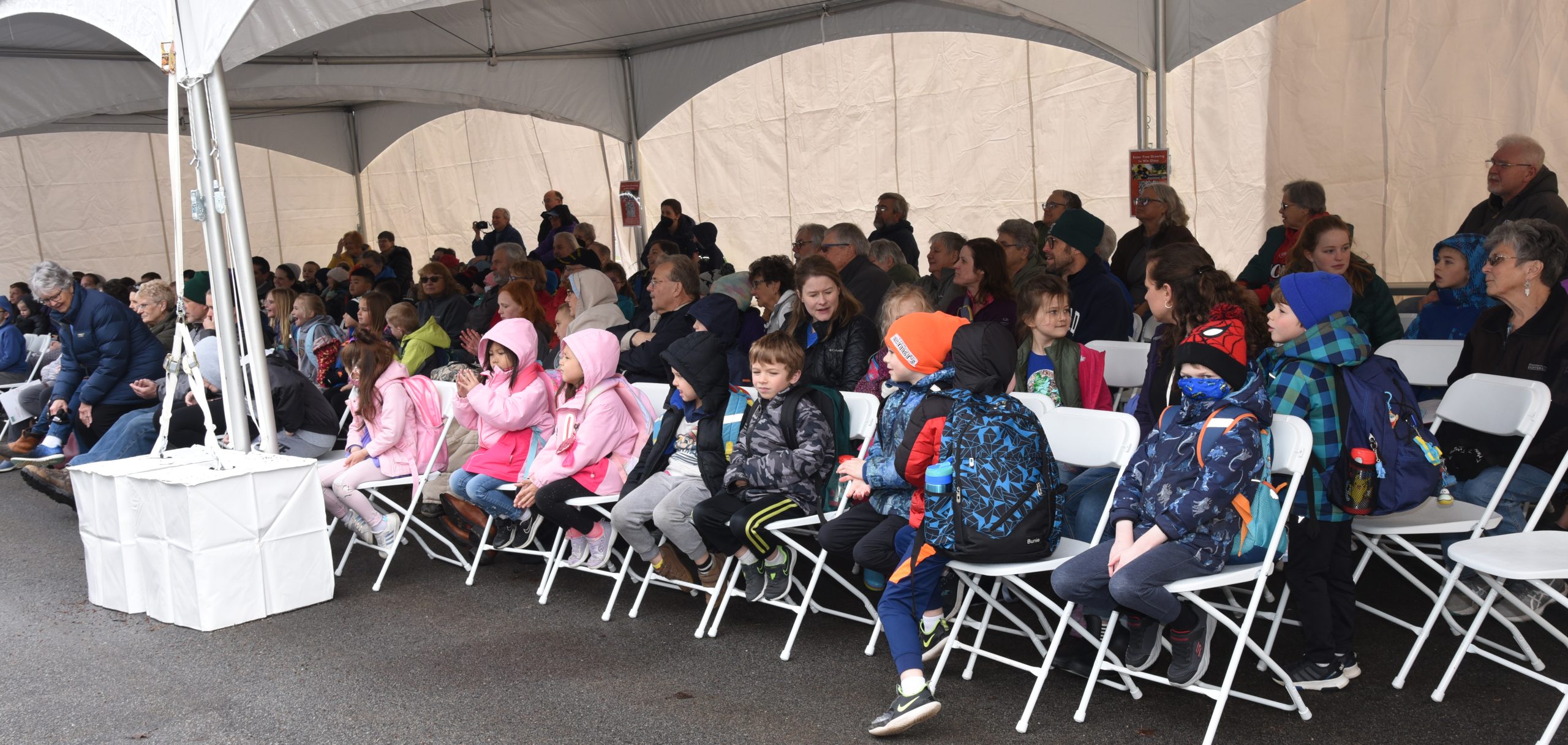Dozens of adults and young students seated beneath a tent listen and watch to glassmaking demonstrations.