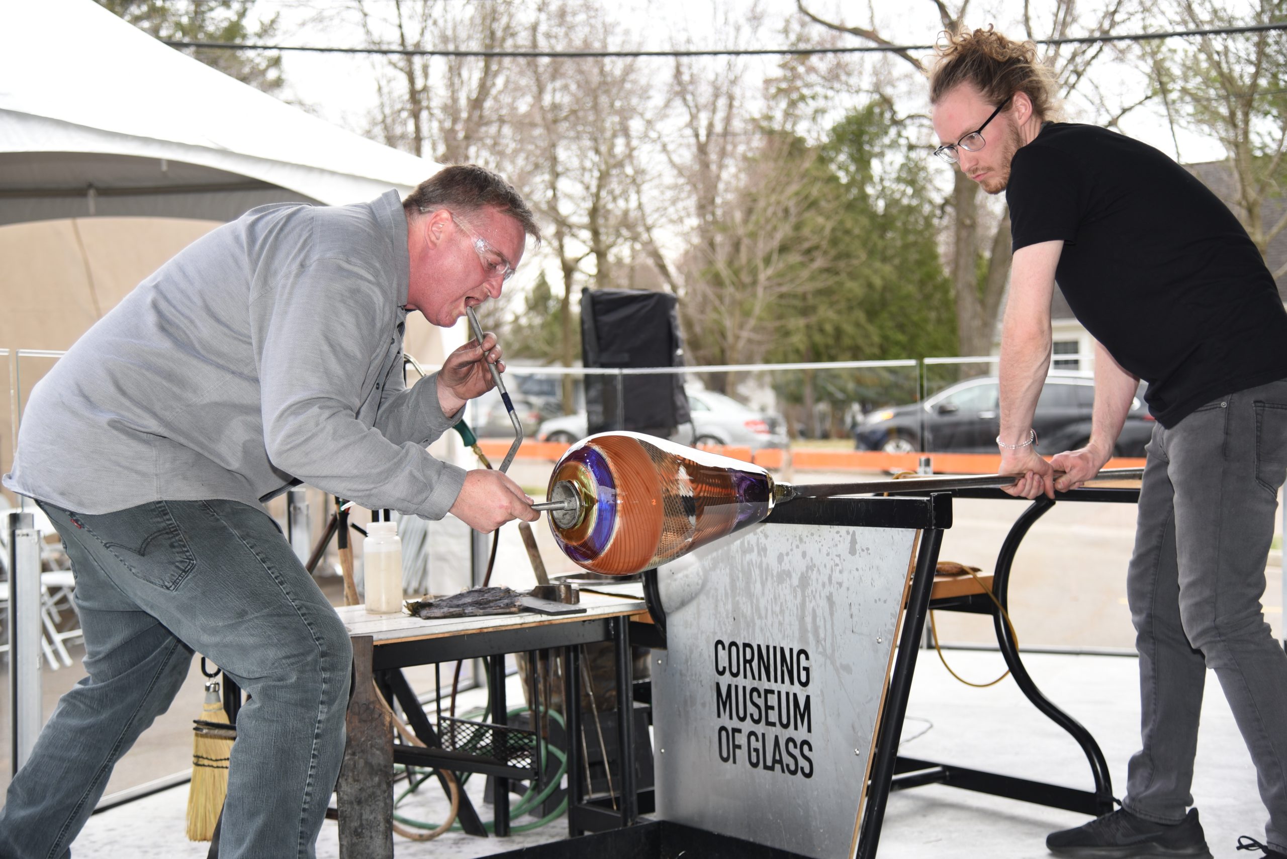 A man crouches forward as he blows air through a tube into colorful molten glass spinning on the end of a metal rod held by another glassmaker.