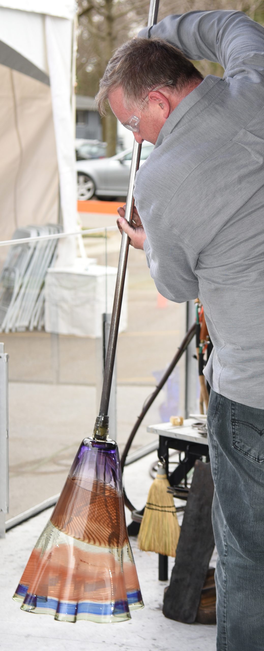 A glassmaker spins a long metal rod that he holds vertically to use gravity to help shape colorfully striped glass while it remains warm and pliable and before it cools and hardens into a large vase.