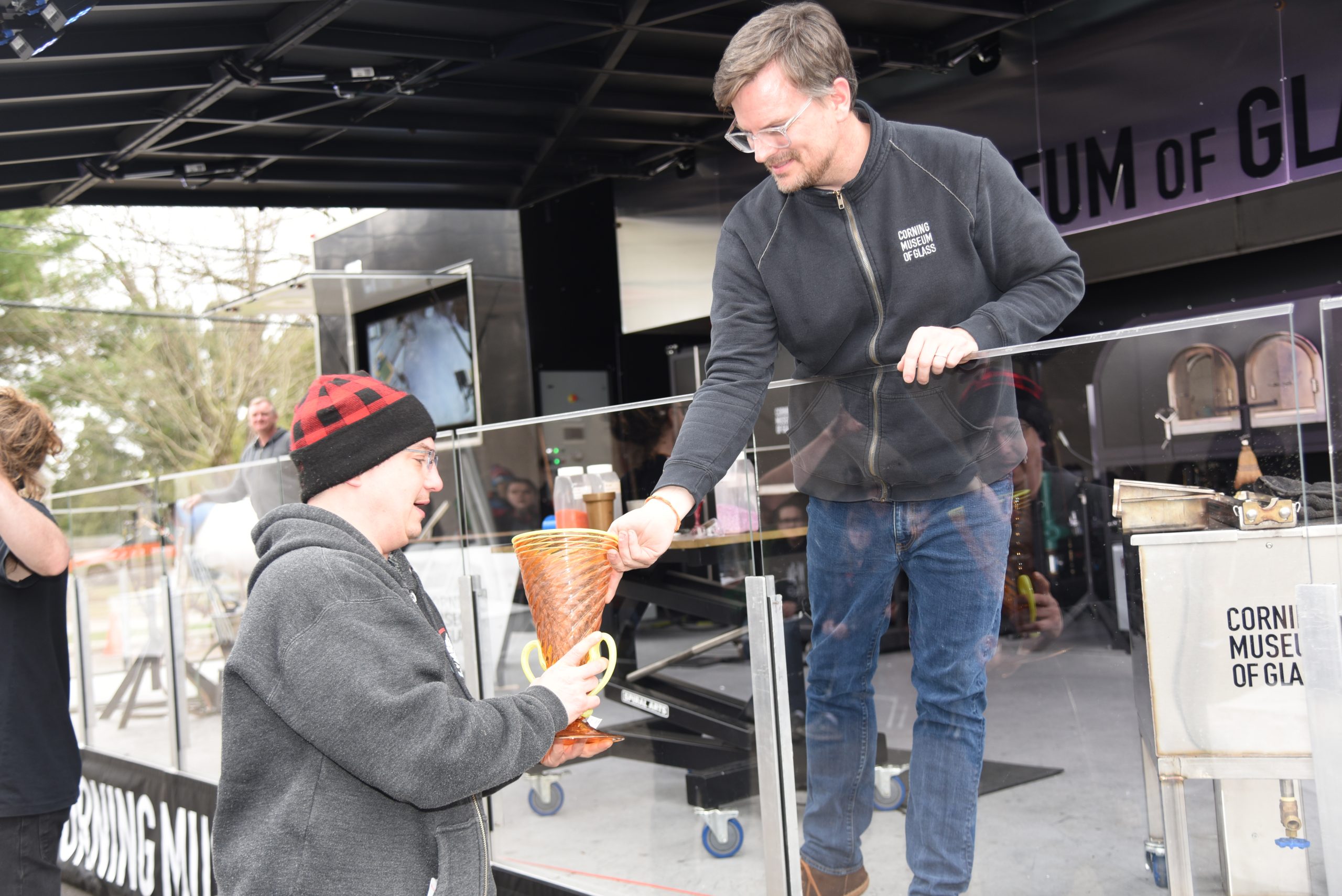 A glassmaker standing on the Mobile Hot Shop stage hands an orange glass vase to a man after a glassmaking demonstration.