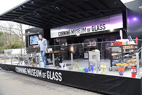 black stage with presenter in middle, labeled Corning Museum of Glass, numerous glass vases and bowls line the stage