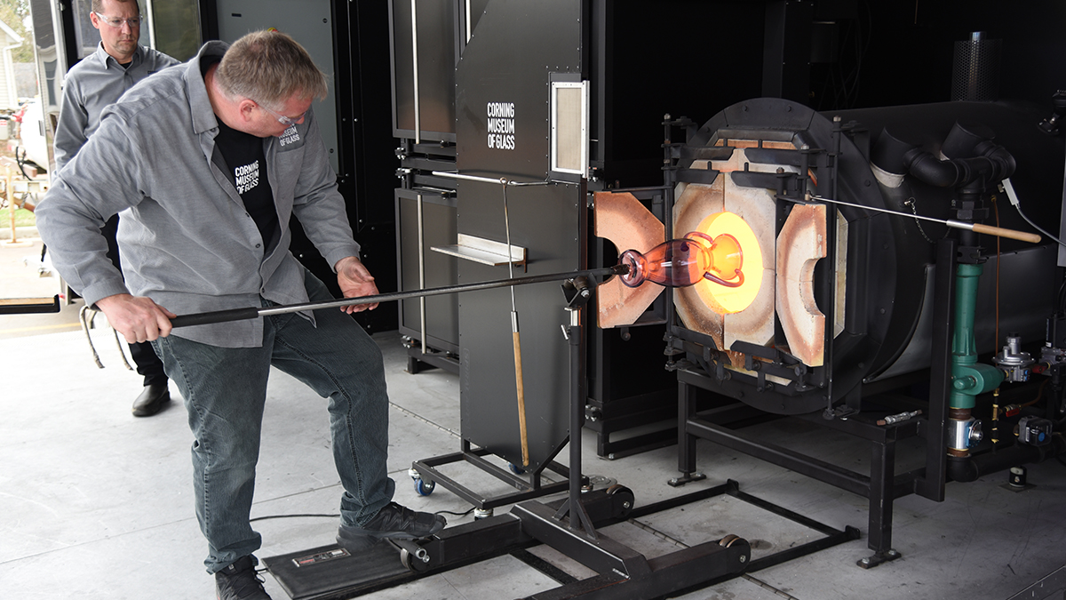 A glassmaker holding a long metal rod pushes a large orange glass vase into a 2,000-degree glassmaking furnace, glowing bright yellow from the heat.