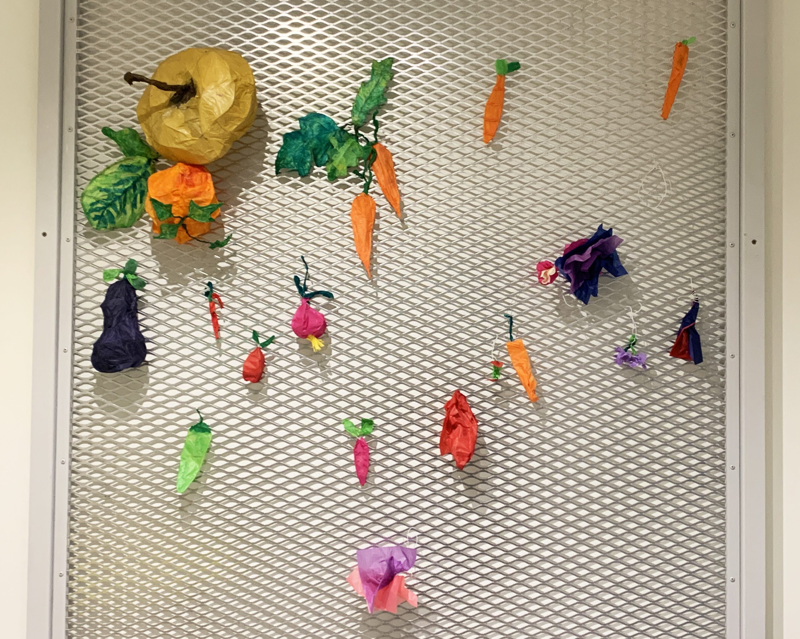 Tissue paper sculptures of carrots, eggplants, and pumpkins made up of various colors, shapes, and sizes hang on the community garden wall in Art Park.