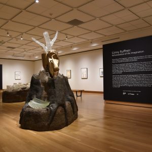 Sculptures of landmasses are dispersed throughout the gallery. Glass stumps with tree rings sit atop each landmass. The surrounding walls are filled with framed drawings of invented plant species.