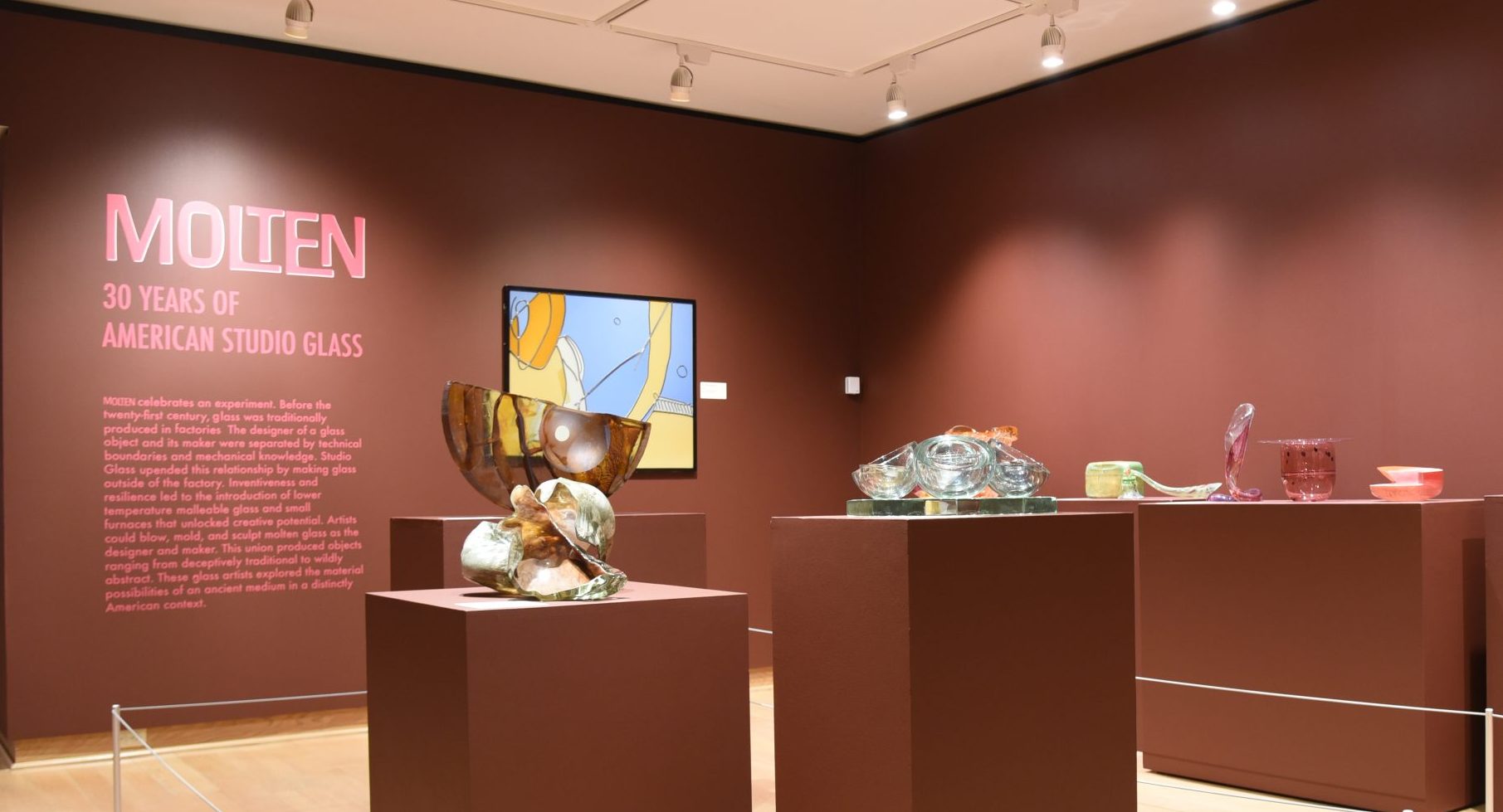This image shows several abstract works of glass on pedestals in the foreground with the Molten exhibition text panel in the background