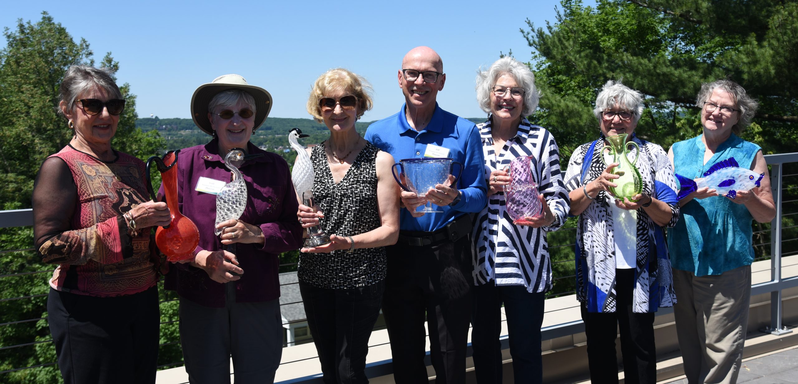 Seven people smile as they each hold a glass sculpture gift on a sunny day with blue sky and trees in the background.