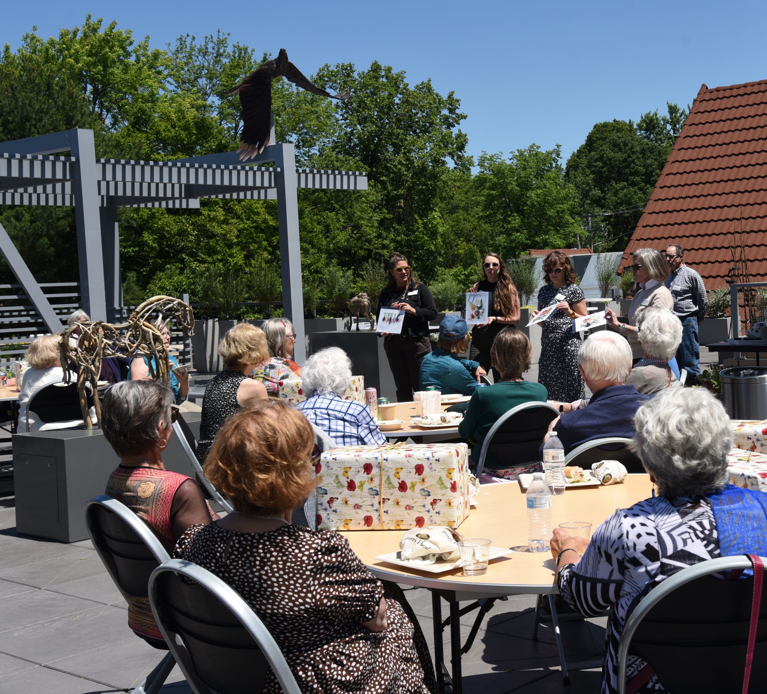 Four people hold prints of colorful fruits and vegetables so docents gathered at tables can see the variety of gifts they received during a volunteer-appreciation luncheon.