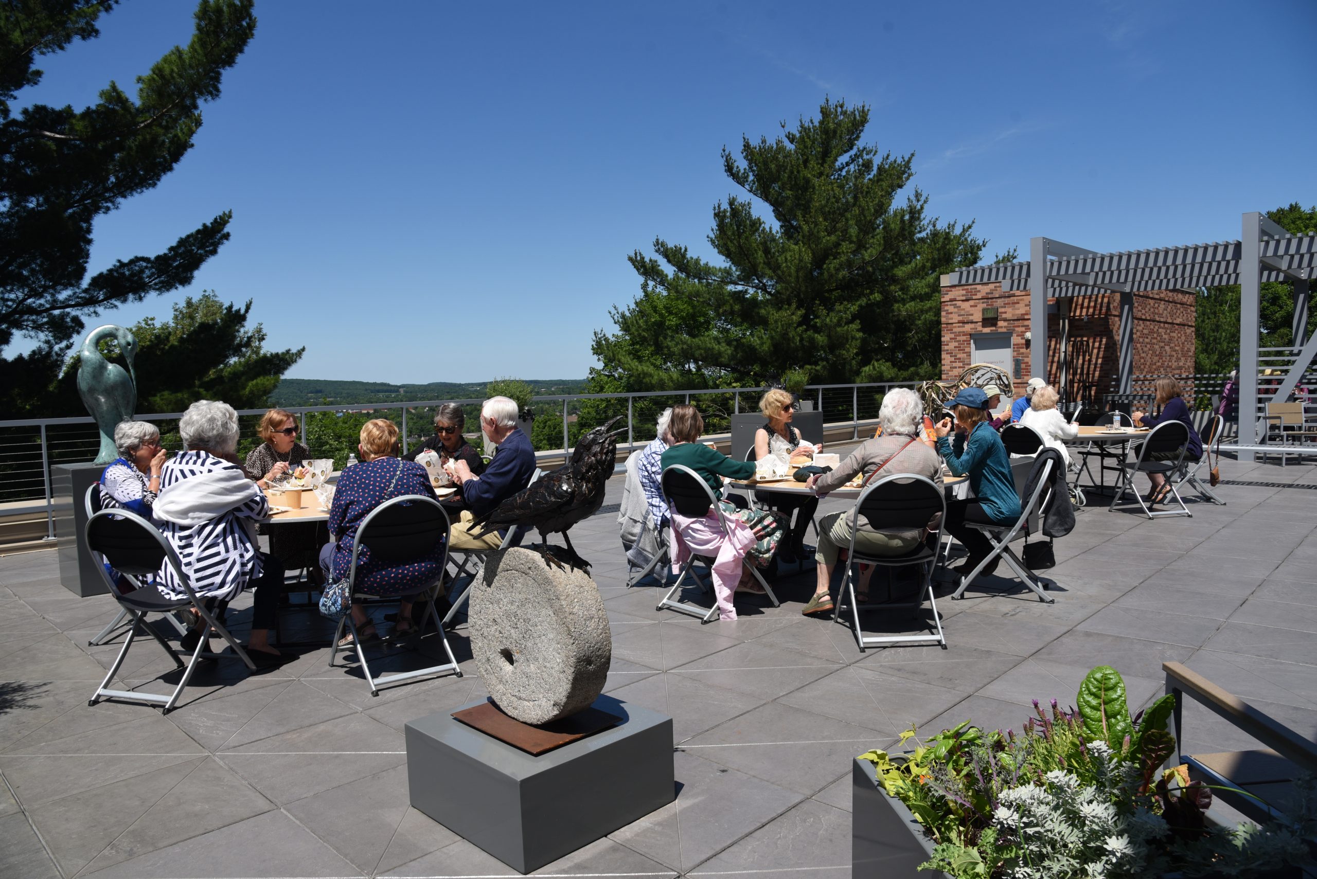 People converse during lunch, seated at tables in a rooftop sculpture garden on a sunny day with blue sky and trees in the background.