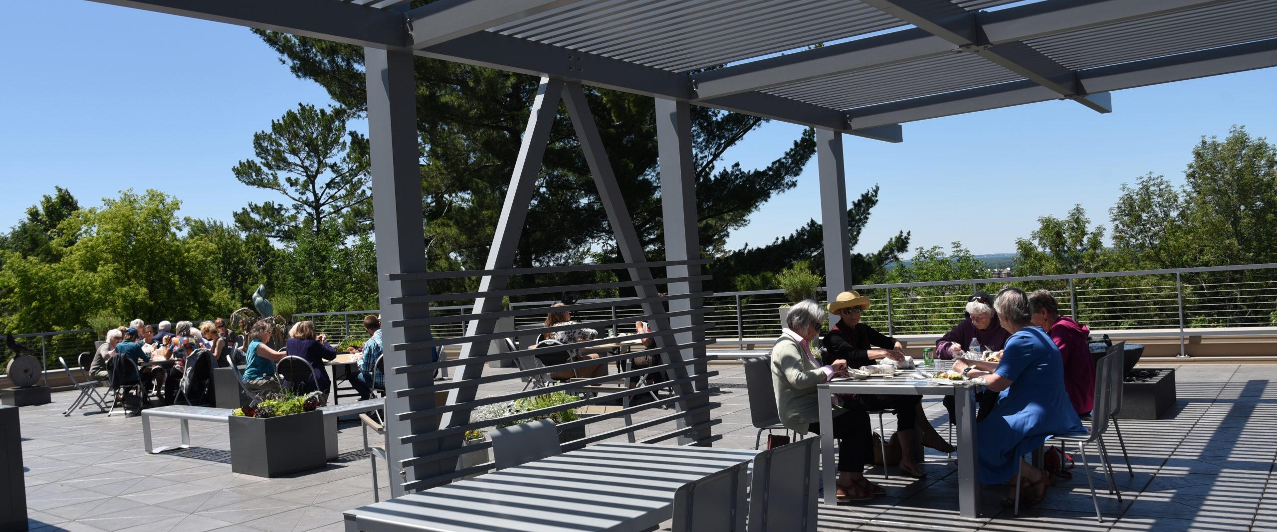 People enjoy lunch, seated at four tables in a rooftop sculpture garden on a sunny day with blue sky and trees in the background.