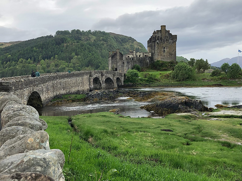 A Scottish castle of gray stone is surrounded by green grass and trees.