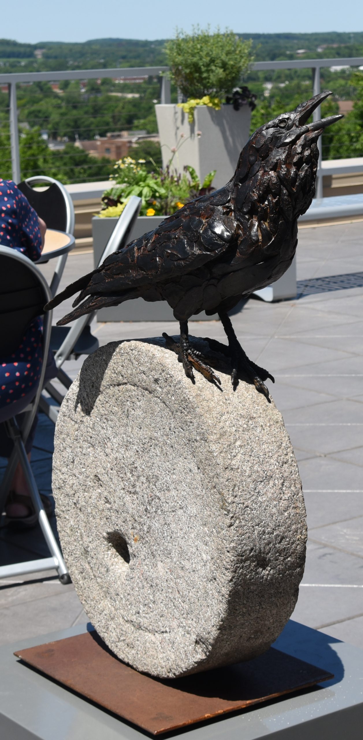 A dark brown bronze sculpture of a raven with head raised and beak open stands atop a circular stone.