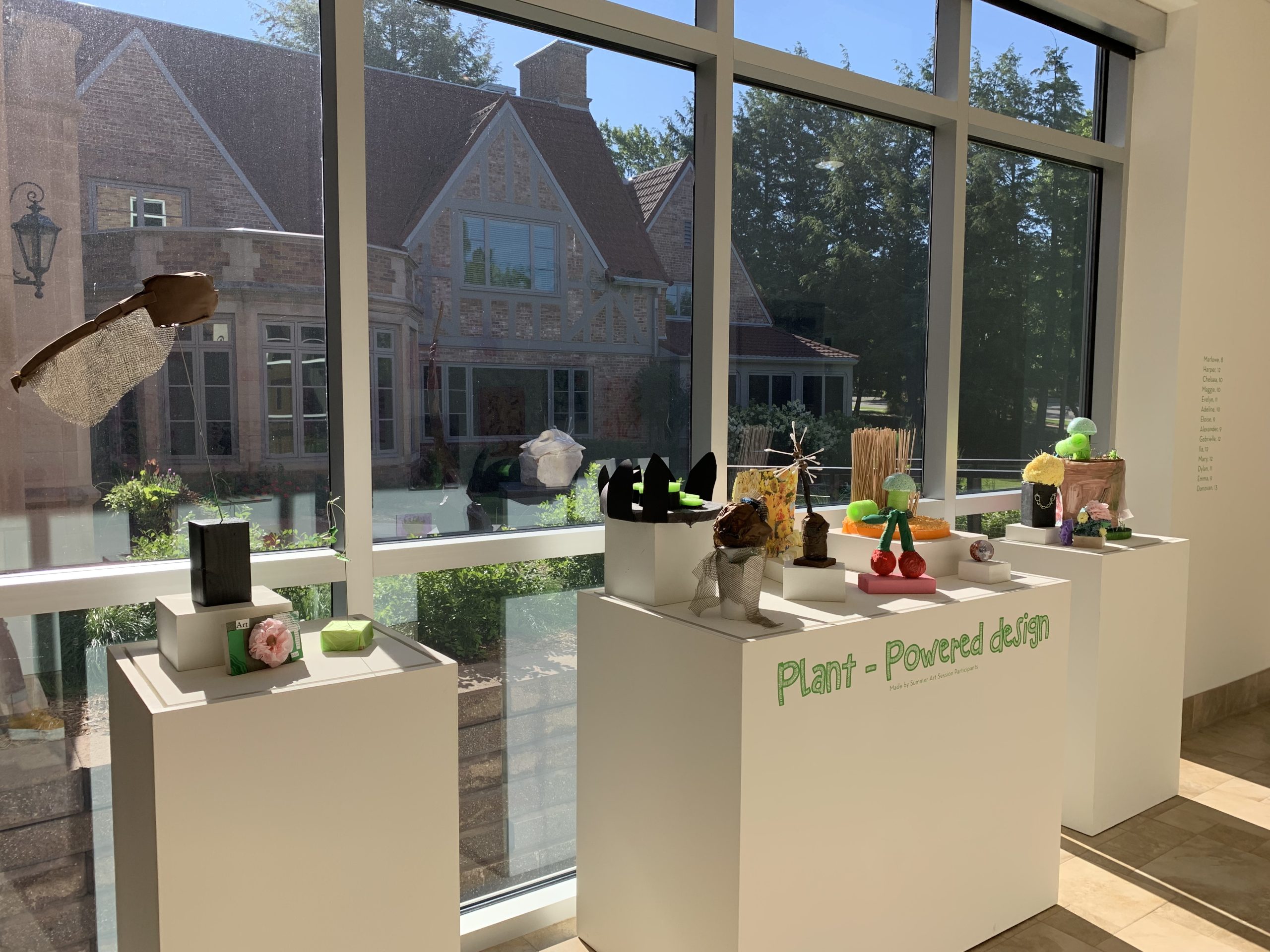 This is a second image of the Art Session projects display, featuring the three-dimensional sculptures created by older students. The projects are displayed in small groups on top of a three white pedestals, shaped like rectangles, positioned in front of a sunny window; outside the window is the pathway to the Museum's main entrance doors where you can see part of the Museum building and garden beds.
