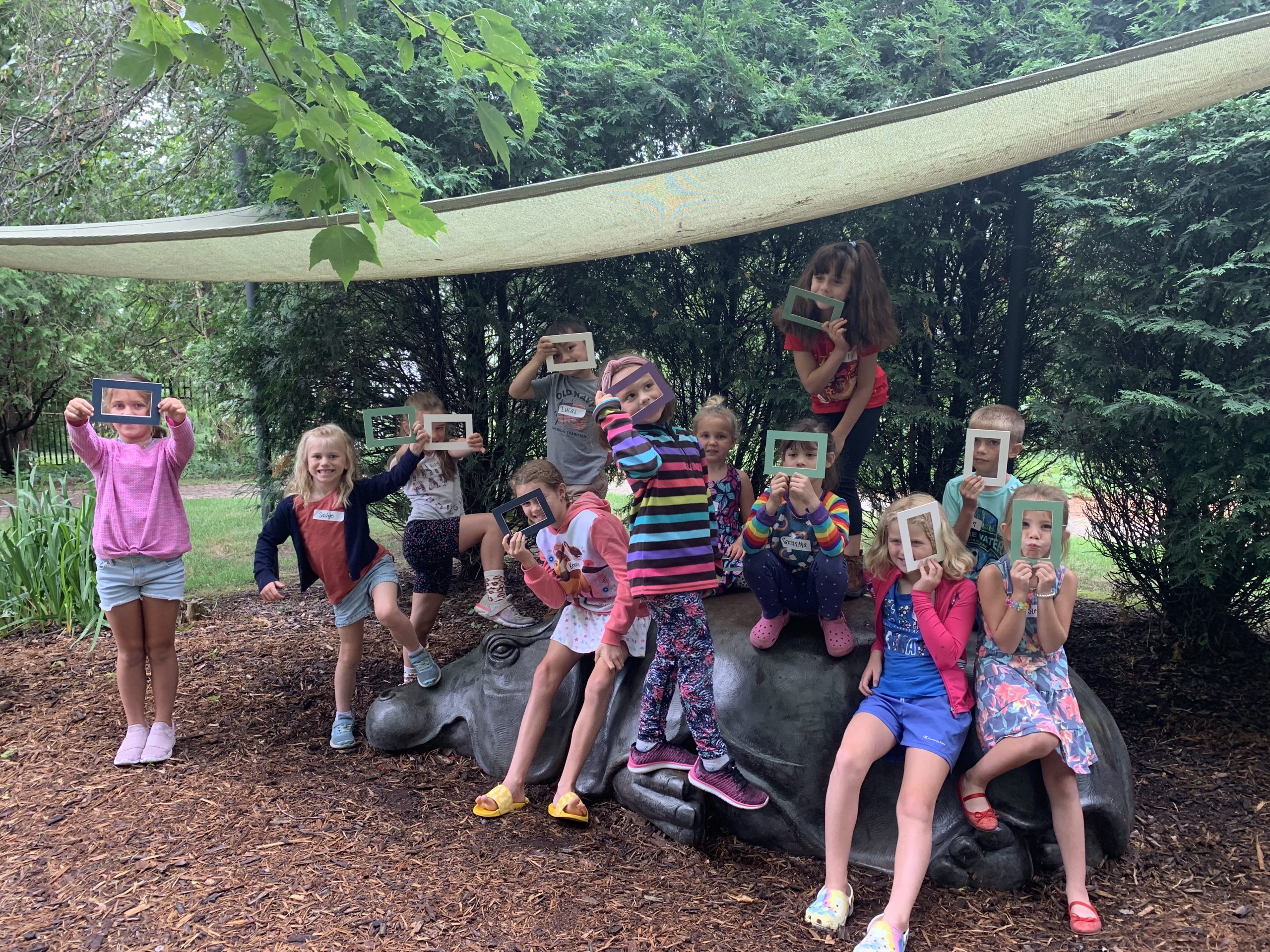 Younger Summer Art Session participants smile and pose behind mat board "view finders" for a group photo, posed around and sitting on top of a bronze hippo sculpture in the garden.