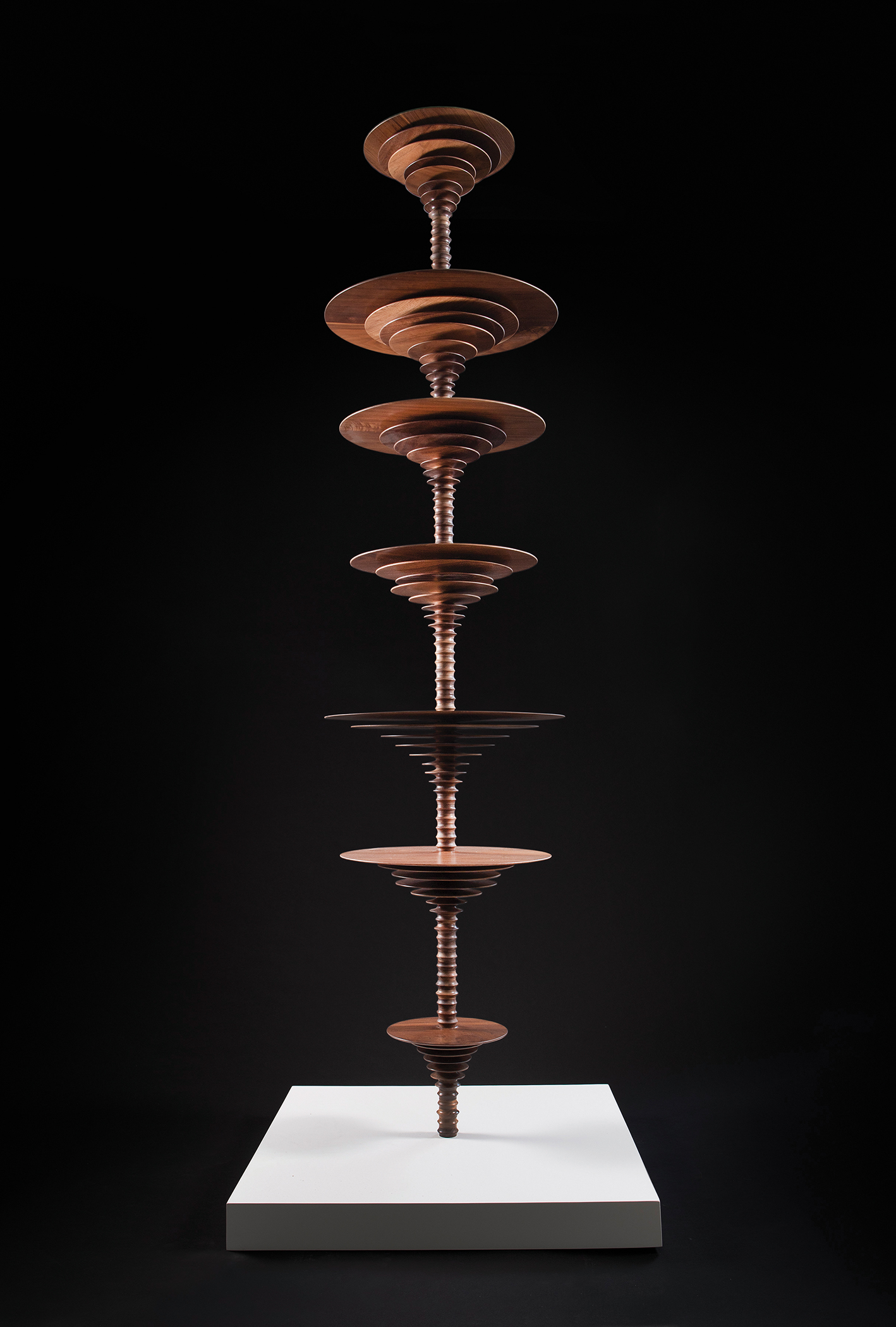 Seven wooden discs of varying sizes are arranged vertically on a wooden column, resting on a white square base