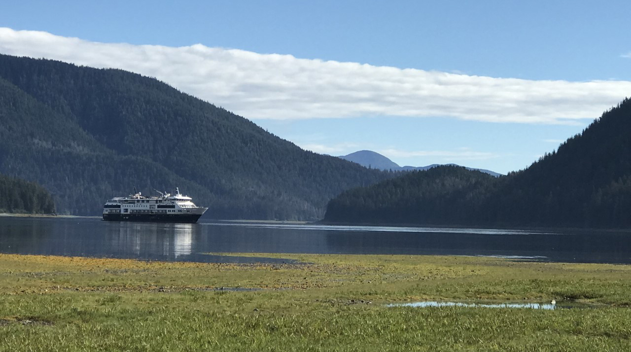 A cruise ship is on calm, smooth water with tree-covered mountains in the background and a blue sky with a strip of white clouds above.