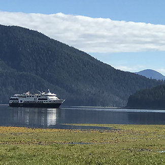 A cruise ship is on calm, smooth water with tree-covered mountains in the background and a blue sky with a strip of white clouds above.