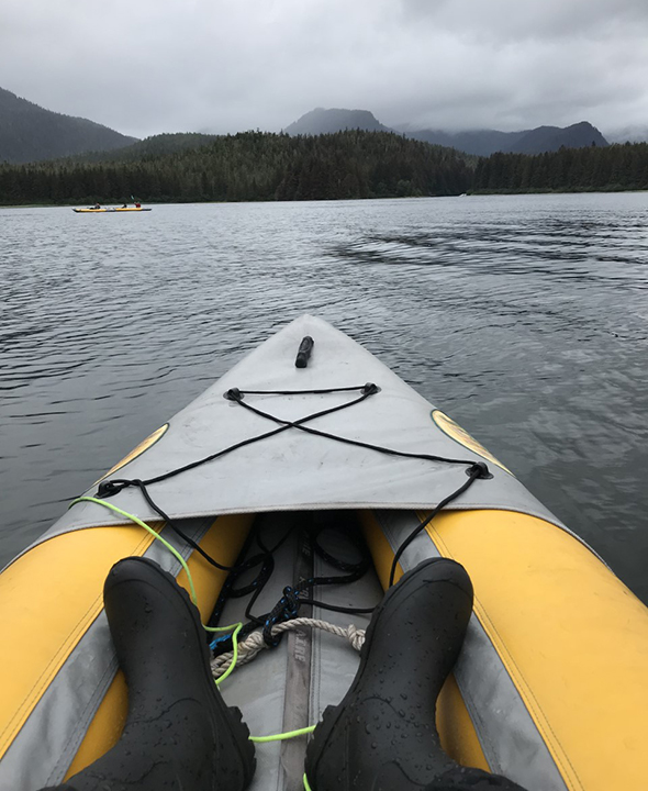 Two black rain boots are near the front of a yellow and gray kayak gliding across dark gray water toward a tree-covered, hilly shoreline with mountains in the distance and a cloudy gray sky above.