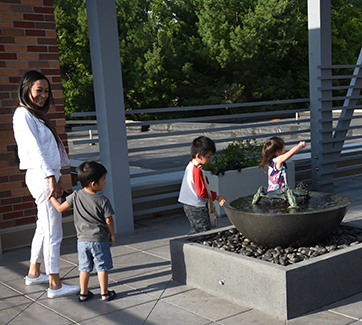A smiling woman and three children are near a rooftop fountain with water-filled bowl and sculpture of frogs and lily pad