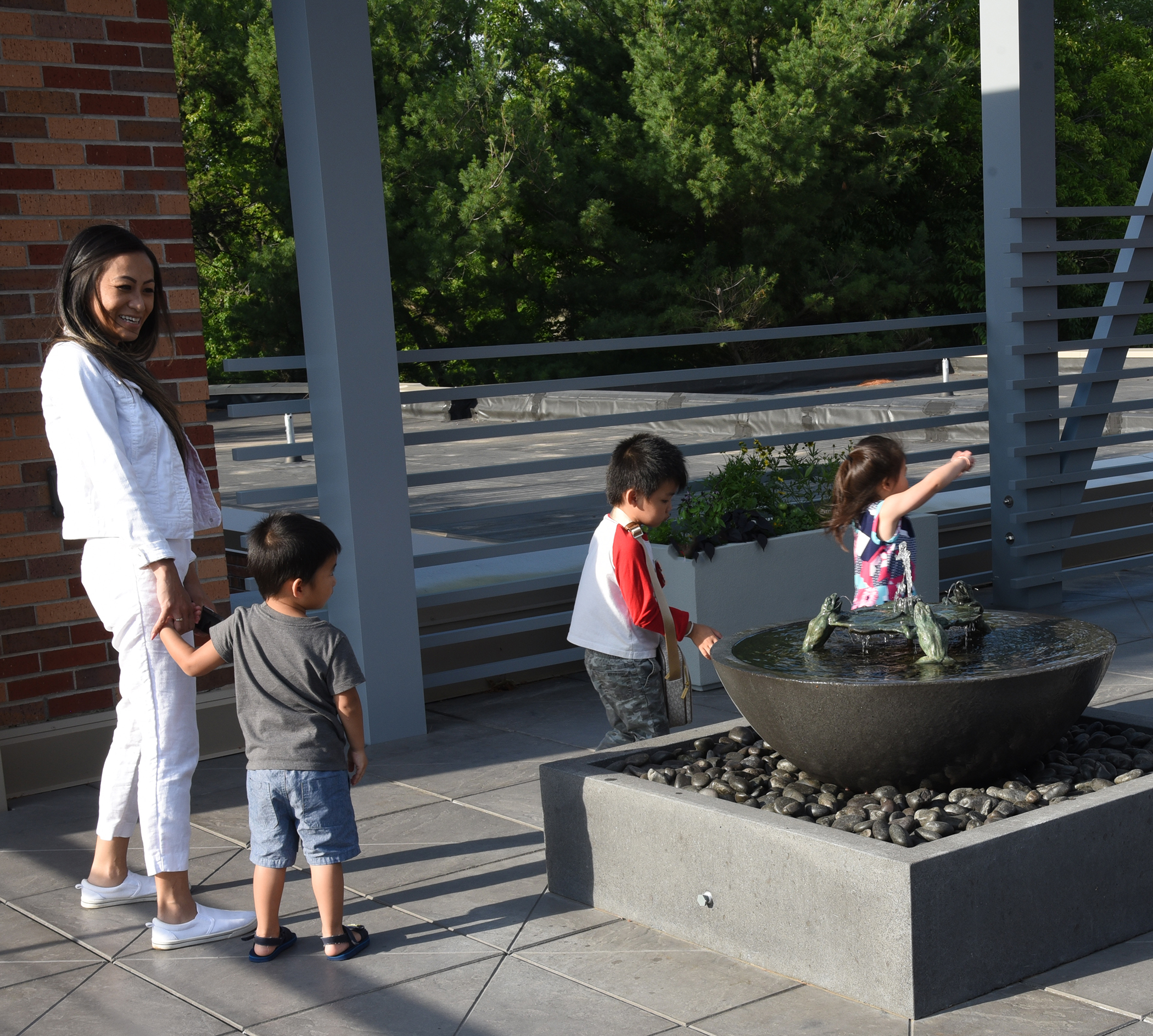 Three young children and a smiling woman gather near a fountain, featuring a large water-filled bowl with a sculpture of four frogs and a lily pad