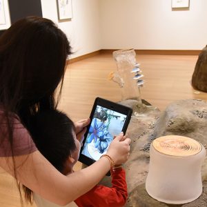 A woman leans over a young child as they hold an iPad and look at the screen that shows a daisy-like flower with blue petals that seems to sprout from glass tree-stump sculptures in a museum gallery.
