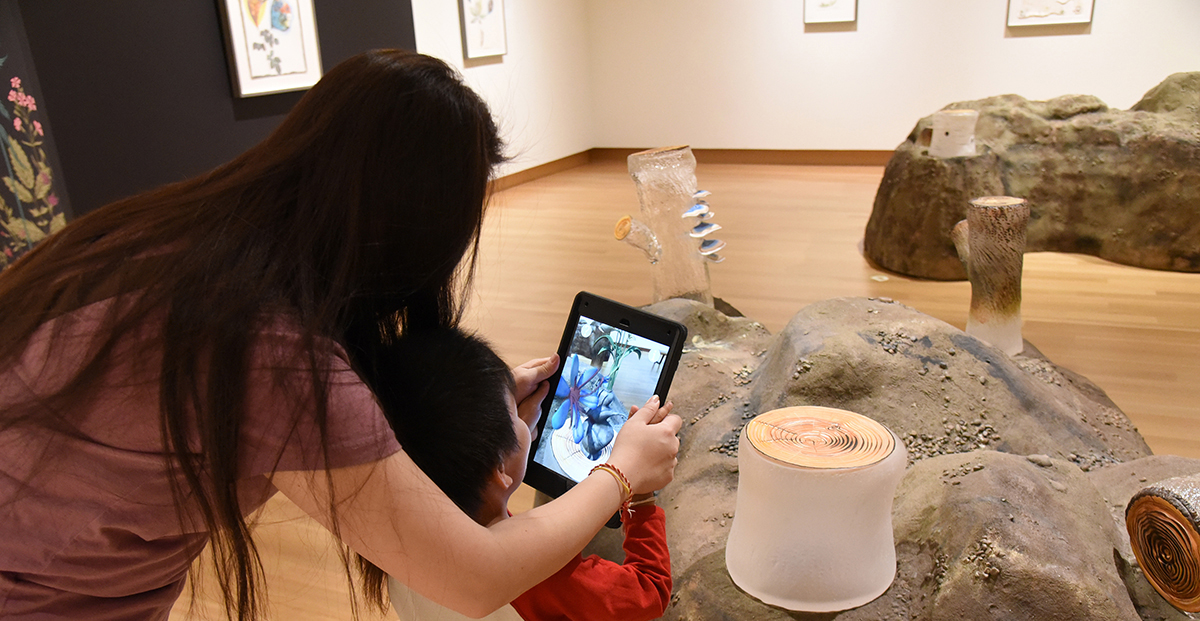 A woman leans over a young child as they hold an iPad and look at the screen that shows a daisy-like flower with blue petals that seems to sprout from glass tree-stump sculptures in a museum gallery.
