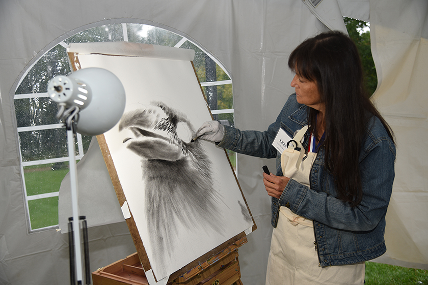 On the left, is a drawing of a black bird, on the right is a woman adding to the drawing with a piece of charcoal