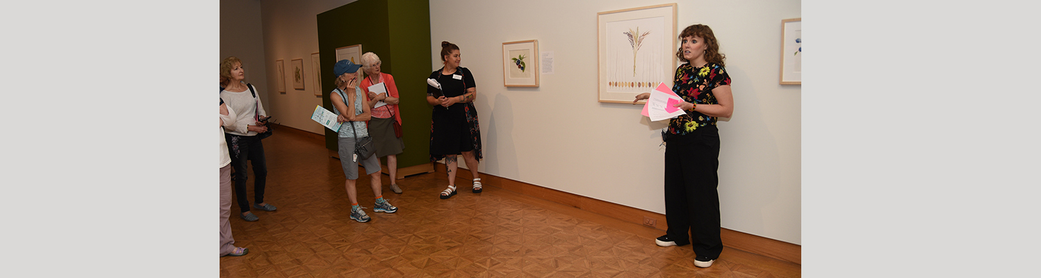 Two museum educators stand near artwork in the galleries talking with volunteers during docent training.