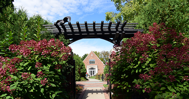 Woodson Art Museum main entrance in the distance, framed by the sculpture garden gate surrounded by pink blooming flowers.