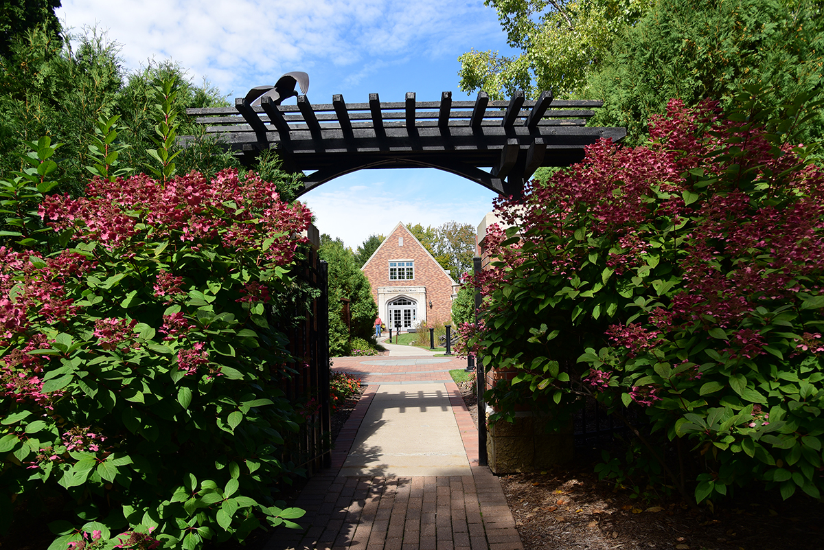 Woodson Art Museum main entrance in the distance, framed by the sculpture garden gate surrounded by pink blooming flowers.