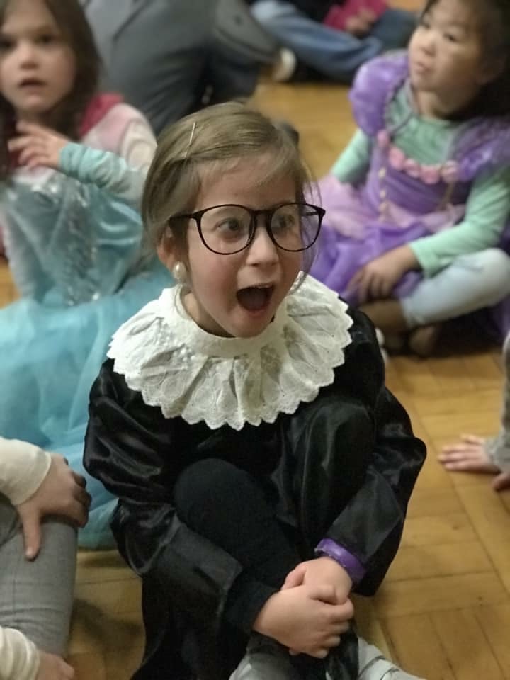 This photo shows a young girl dressed up as Supreme Court Justice Ruth Bader Ginsburg complete with glasses, earrings, and a black robe. She is sitting on a floor surrounded by other children in Halloween costumes, mostly wearing princess outfits.