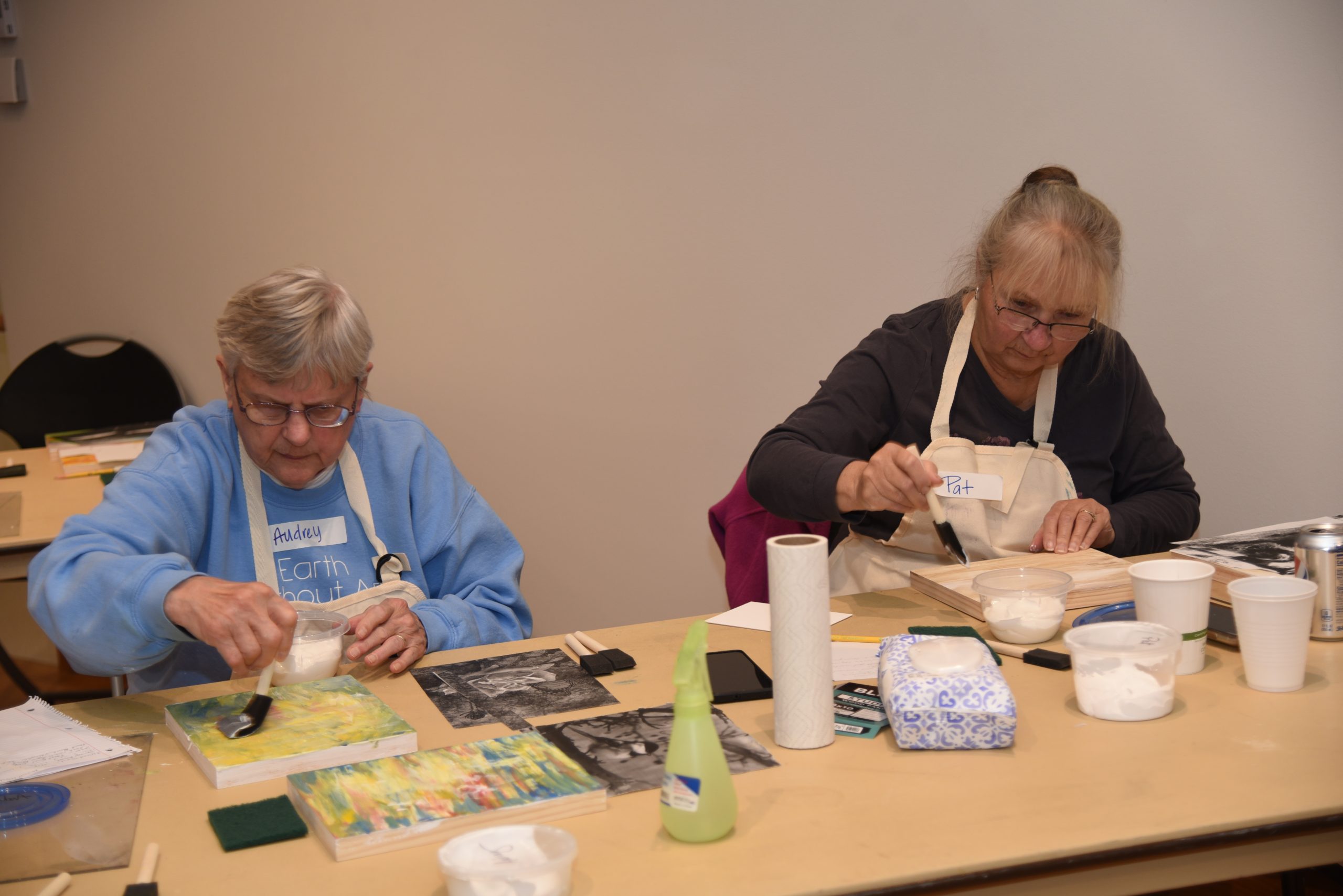 Two workshop participants sit aside one another working on wooden panels. 