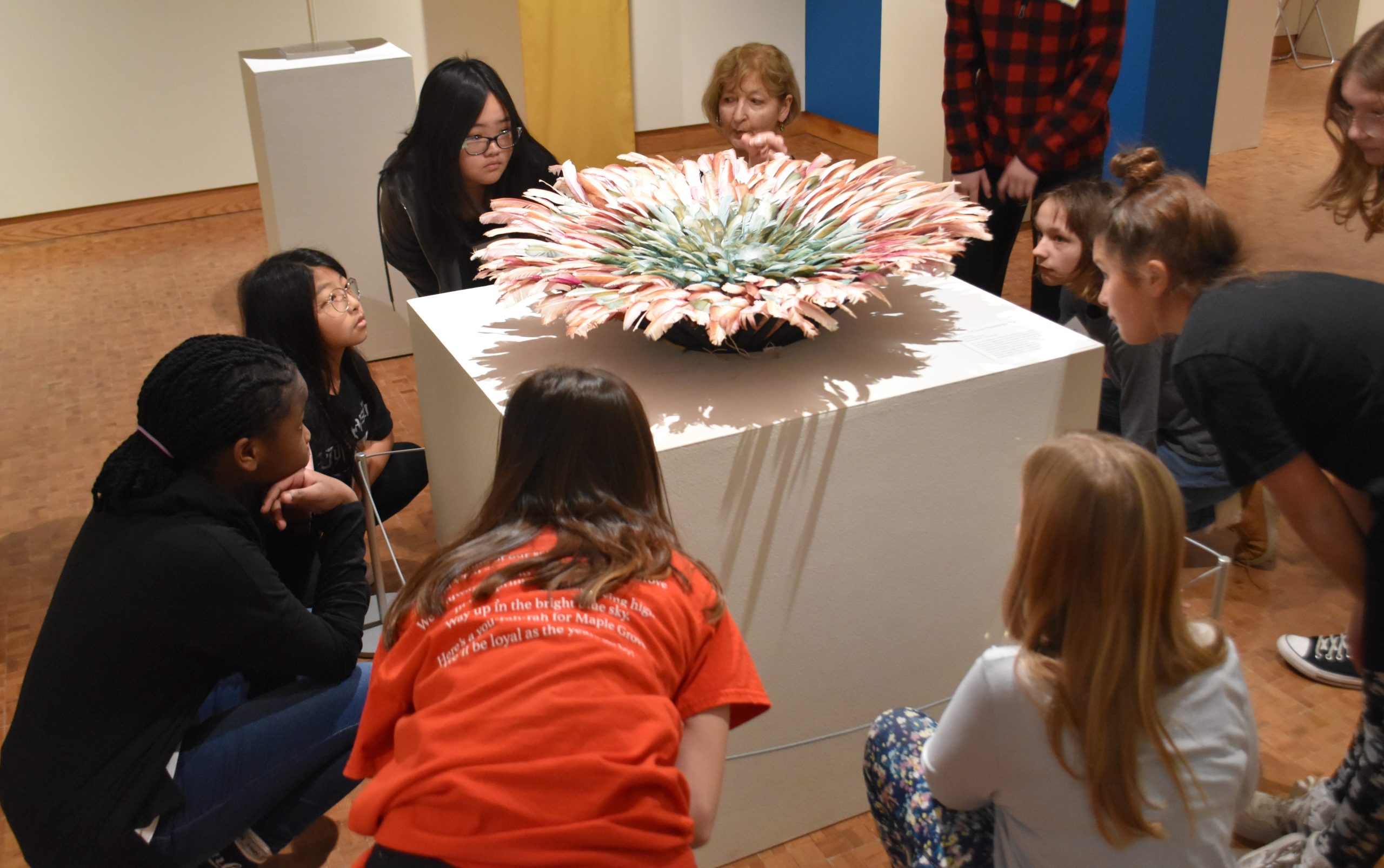 Eight students and volunteer docent crouch down to view a large, round headdress made of feathers on a pedestal in the Museum gallery.