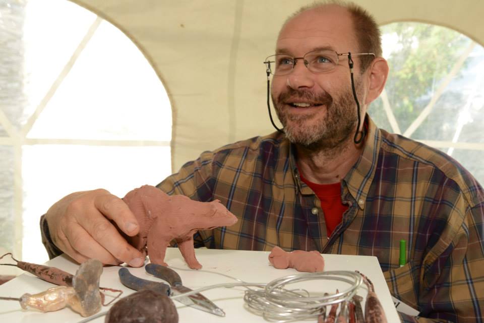 Paul Rhymer is pictured at a table, looking up and smiling at someone off camera, with his right hand holding a small clay bear. Also on the table in front of him are needle-nose pliers, wire, and clay tools used during his sculpting demonstration. 