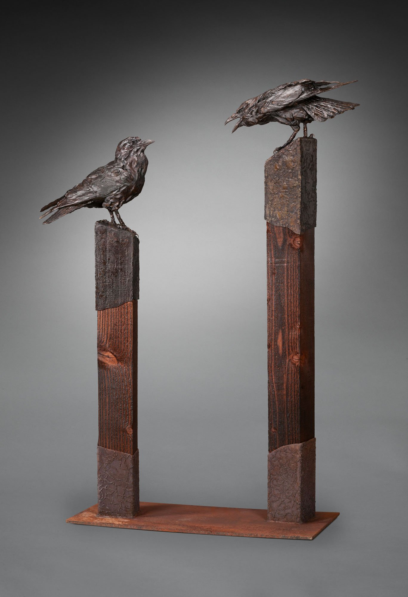 Picture of Paul Rhymer sculpture featuring two tall posts, each with a raven perched on top. The raven on the taller post, at left, is leaning forward toward the second across from it, with its beak open, as if lecturing the second bird. The second raven, at right, on the slightly shorter post is looking at the first bird calling at it.