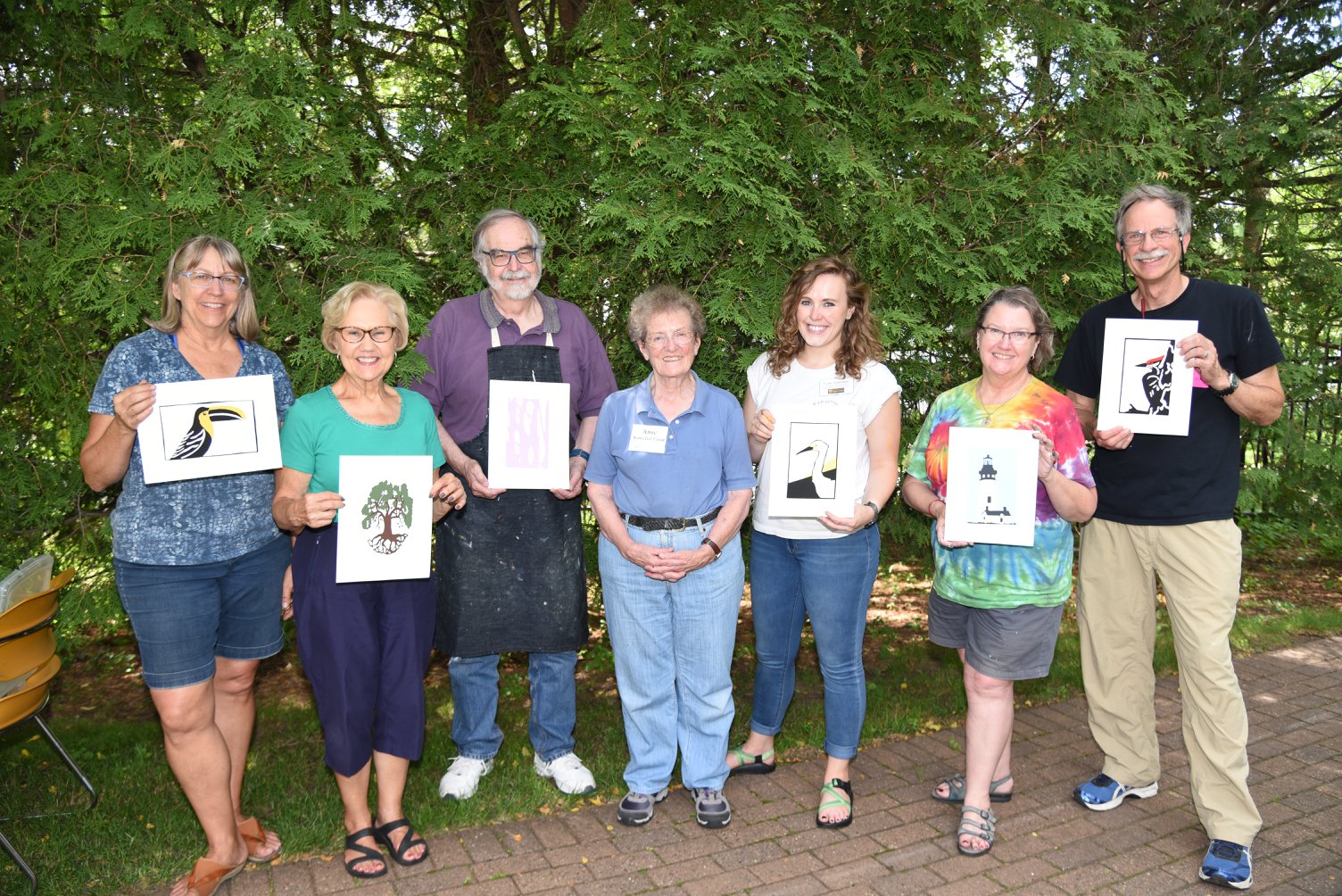 A group photo of seven adults, six participants from a screen printing workshop are holding up examples of their prints while the instructor stands in the middle of the group.