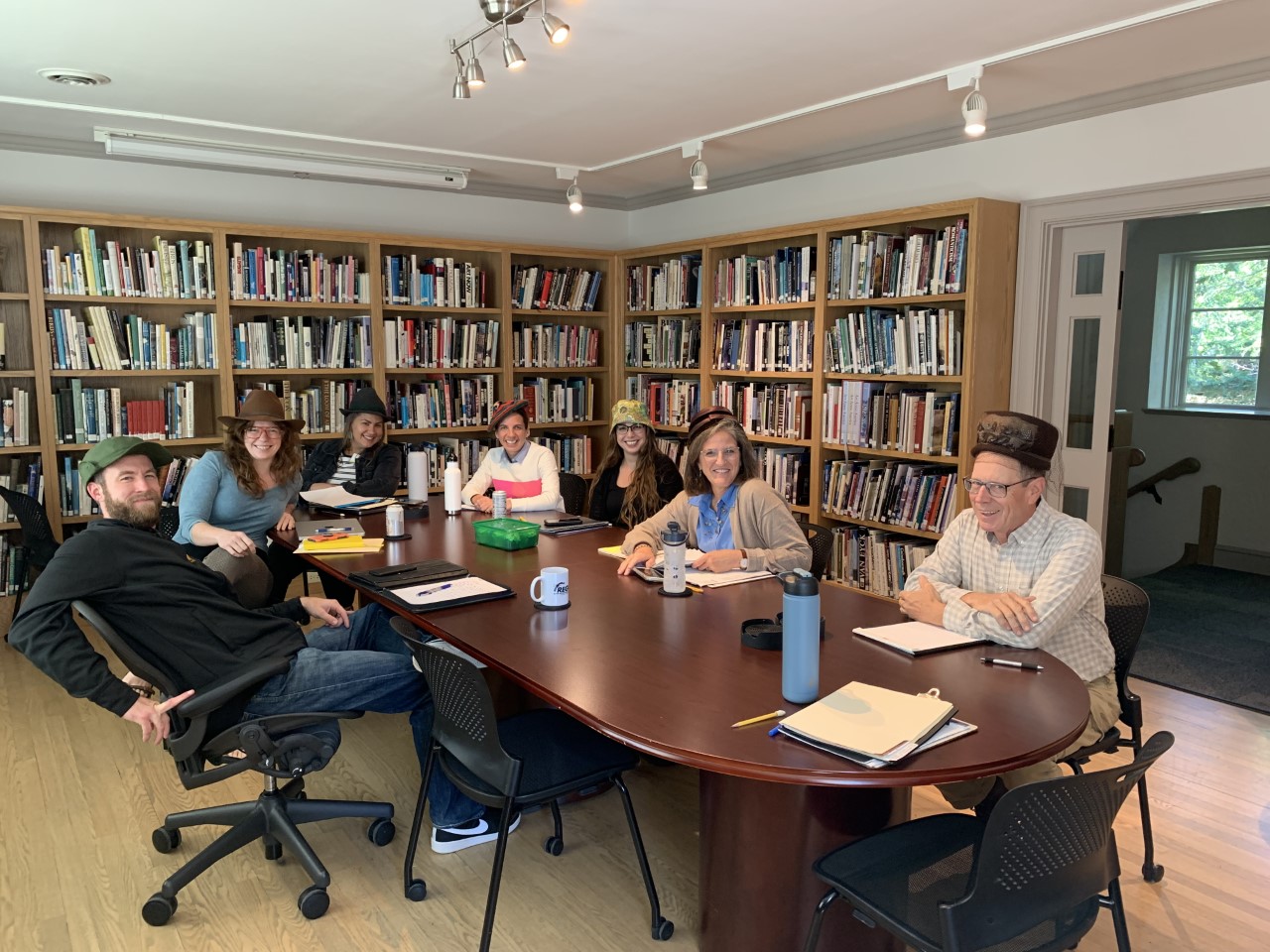 Museum staff members seated around a large conference table in the library, each person has on a different hat.