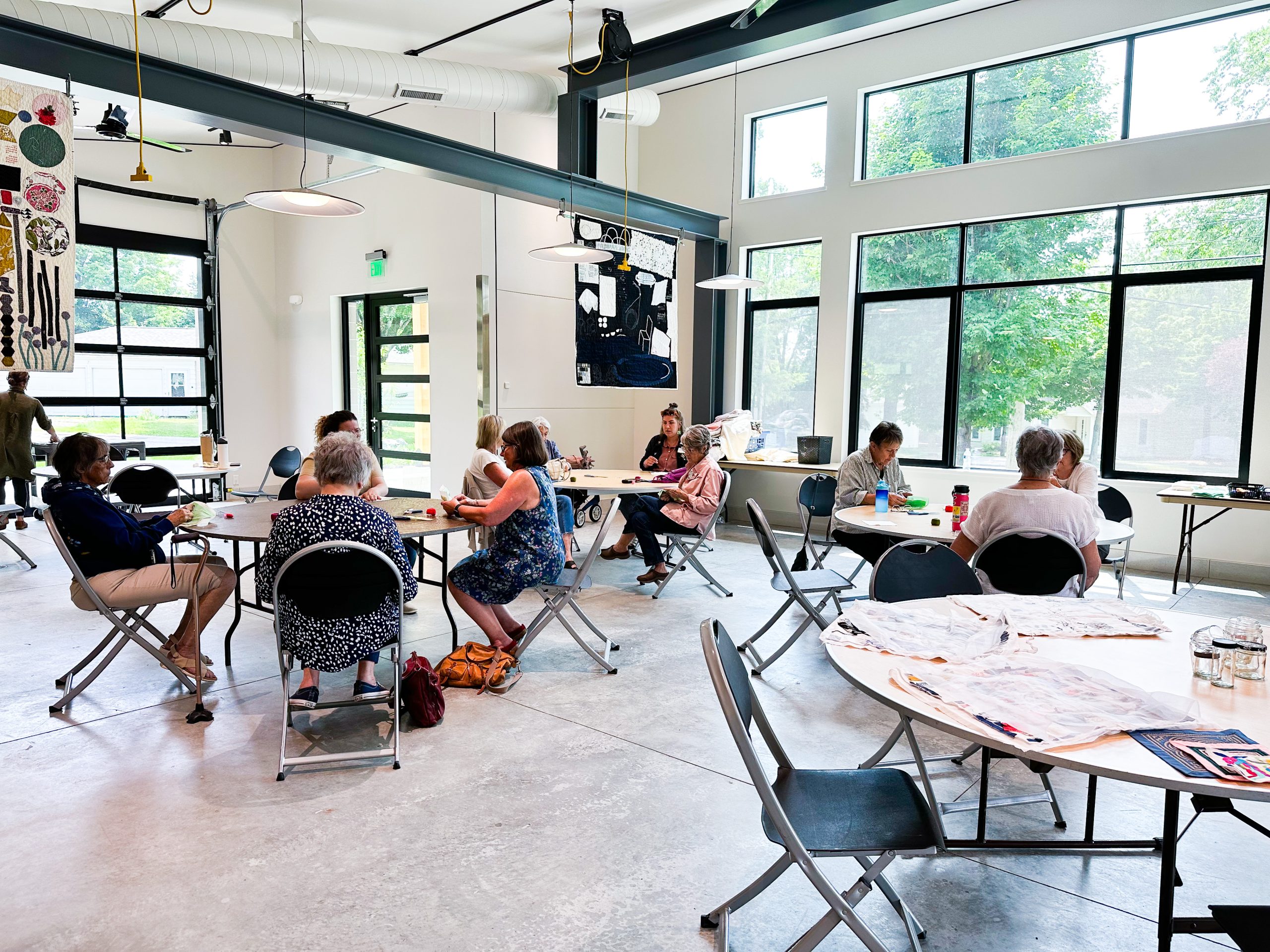 People sit at round tables in a large room with natural light. They are working on small, quilted squares during a drop-in program led by guest artist Heidi Parkes.