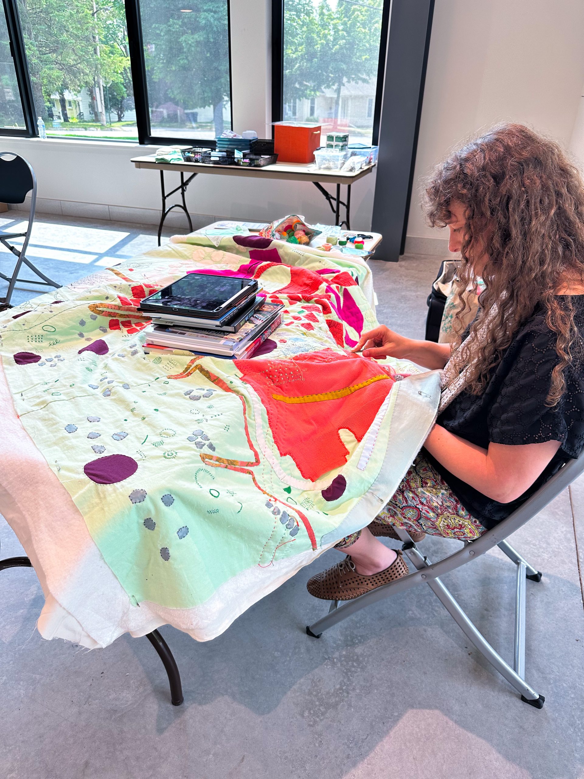 Heidi Parkes sits at a table working on her large, colorful quilt made from green and red-orange fabric.