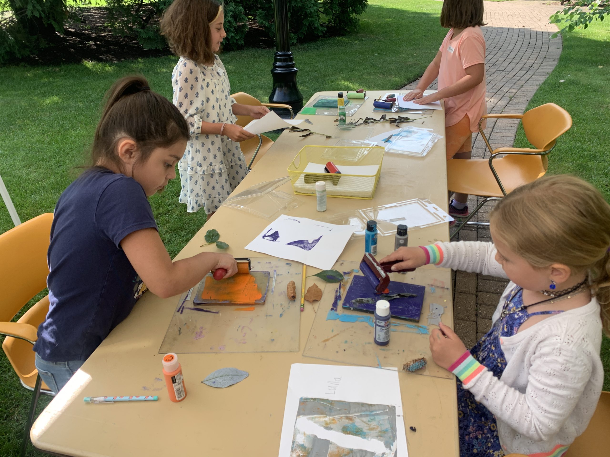 Children are rolling out paint onto Gelli plates to create prints with found materials like leaves, flowers, and sticks.