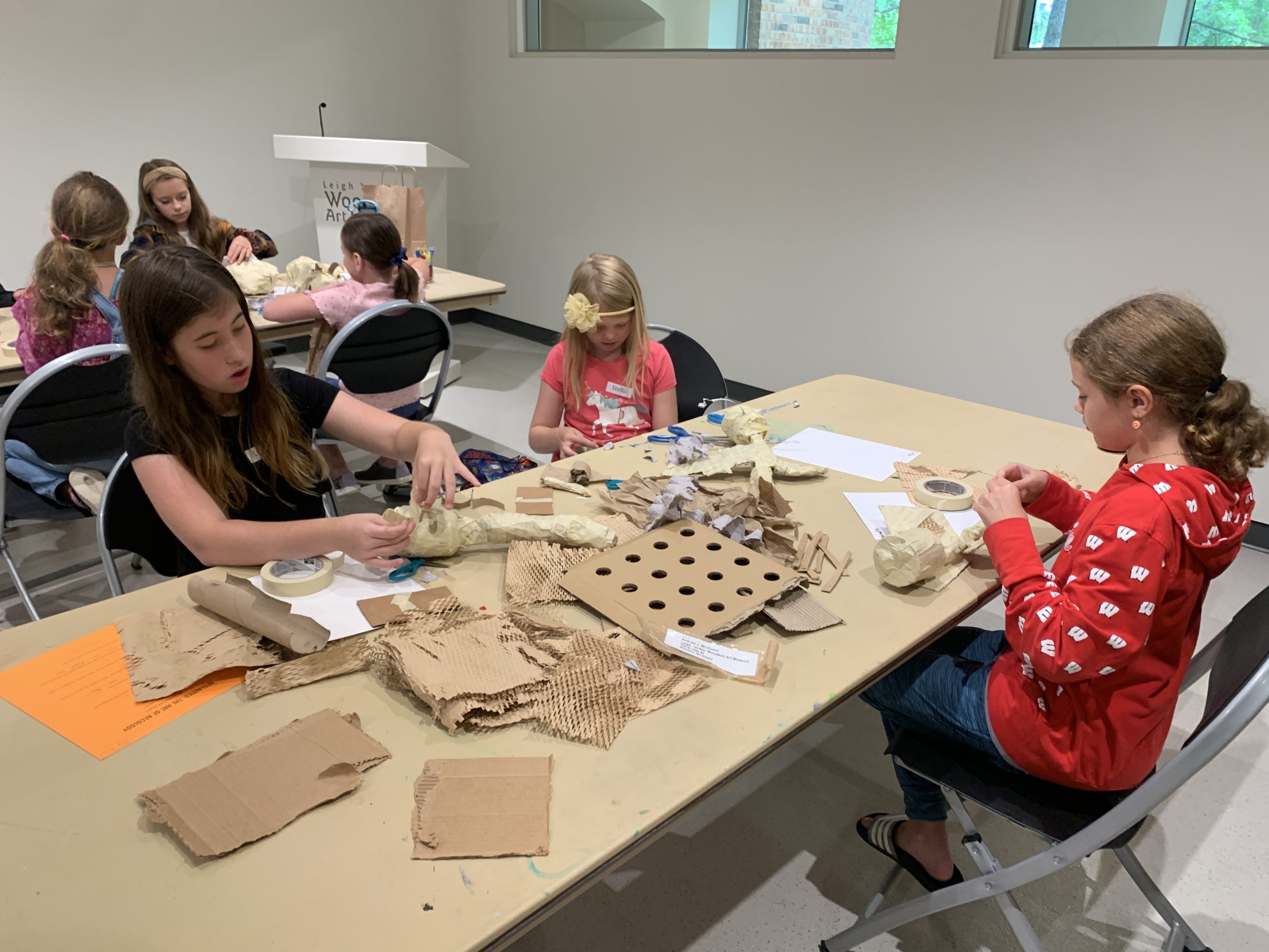 Children sit at a large folding table filled with cardboard, paper, and tape they are using to created recycled insect sculptures. 